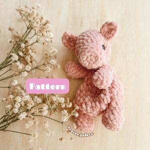 May include: A pink crocheted plush toy shaped like a pig. The toy is lying on a light brown wooden surface with white flowers.