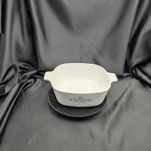 Könnte beinhalten: Eine weiße, quadratische Auflaufform mit zwei Griffen und einem blauen Blumendesign. Die Schüssel steht auf einem kleinen, runden, schwarzen Teller. Der Hintergrund ist ein dunkler, drapierter Stoff.