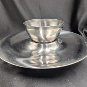 以下が含まれることがあります： 中央に小さなボウルが付いた銀製のサービングディッシュ。ディッシュは丸く、わずかに盛り上がった縁があります。ボウルも丸く、滑らかで光沢のある表面をしています。