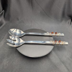 Könnte beinhalten: Ein silbernes Salatbesteck-Set. Die Griffe sind mit braunen Perlen verziert und mit silbernem Draht umwickelt. Das Set liegt auf einem schwarzen Teller vor dunklem Hintergrund.