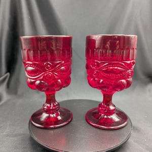 Op de afbeelding: Twee robijnrode vintage kelken met sierlijke, reliëf ontwerpen. De glazen hebben een gedetailleerd patroon rond de kom en een voet. De kelken zijn waarschijnlijk van glas en worden op een donkere ondergrond gepresenteerd.