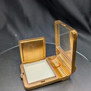 以下が含まれることがあります： ヒンジ付きの蓋と鏡が付いたゴールドトーンの金属製コンパクトケース。内部にはパウダー用の正方形の白いセラミック製インサートと、アプリケーター用の小さなリブ付きのゴールドトーンのセクションがあります。外側の金属面は細かい質感の仕上げになっています。