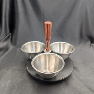 Peut inclure: Un ensemble de service à trois bols avec une poignée en bois foncé et une base noire. Les bols sont en acier inoxydable. La poignée est de couleur marron foncé. L'ensemble est conçu pour servir des sauces ou de petites portions de nourriture.