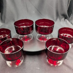 Könnte beinhalten: Sechs rubinrote Dessertschalen mit klaren Glasstielen und silbernem Rand. Die Schalen sind auf einer dunkelgrauen Oberfläche angeordnet. Die Schalen sind tiefrot und scheinen aus Glas zu sein.