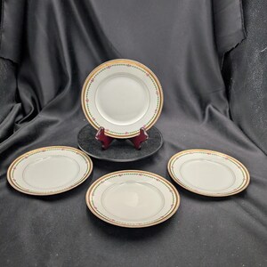 May include: Four white porcelain plates with a gold rim and a floral pattern. One plate is displayed on a black stand. The plates are arranged on a black surface.