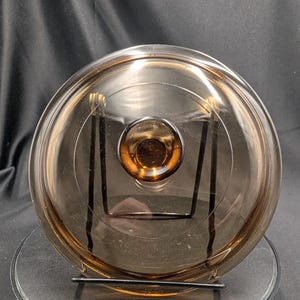 May include: A circular, transparent amber-coloured glass lid featuring a smooth, domed surface and a central recessed knob handle. The glass has a warm, brownish tint and a polished finish, reflecting light across its curved, reflective surface.