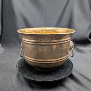 May include: A vintage metal ice bucket with a brass rim and lion head handles. The bucket has a tarnished finish and sits on a black coaster. The design is reminiscent of antique barware.