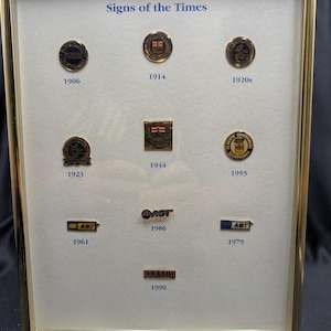 Può includere: Esposizione incorniciata di spille e distintivi vintage, intitolata "Signs of the Times". Il display include spille del 1906, 1914, 1920, 1923, 1944, 1955, 1961, 1979, 1986 e 1990. Le spille presentano vari disegni e testi.