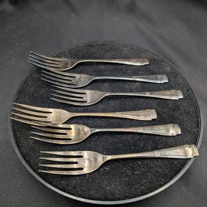 May include: Six silver forks displayed on a black, circular surface. The forks have tines and decorative handles. The forks are arranged in a fan-like pattern, with the handles pointing outwards. The forks are made of a shiny, metallic material.