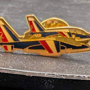 Könnte beinhalten: Goldfarbene Flugzeugnadel mit roten, weißen und blauen Akzenten. Die Nadel zeigt ein detailliertes Design eines Düsenflugzeugs, mit der Nummer 2 auf dem Heck. Die Nadel ist wahrscheinlich ein Sammler- oder Dekorationsartikel.