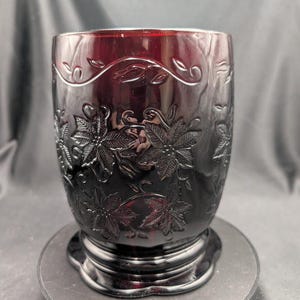Puede incluir: Un vaso de vidrio rojo oscuro con un diseño floral en relieve. El diseño presenta hojas y vides en relieve, creando una superficie texturizada. El vaso se apoya sobre una base circular, mostrando un diseño clásico y elegante.