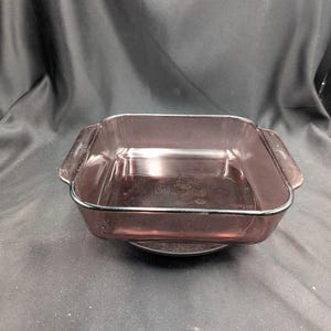 Puede incluir: Una fuente de horno cuadrada de vidrio púrpura transparente con dos asas. La fuente tiene un borde negro alrededor de los bordes y está sobre una superficie oscura. La fuente está vacía.