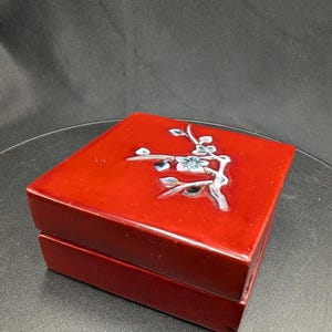 Può includere: Una scatola quadrata in lacca rossa lucida con un intarsio decorativo raffigurante un ramo fiorito sul coperchio. Il ramo è realizzato in tonalità di bianco, grigio e blu, con delicati fiori e boccioli sulla superficie rossa brillante.