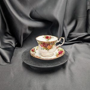 以下が含まれることがあります： 赤、黄、オレンジの色合いの花柄のヴィンテージティーカップとソーサーのセット。 ティーカップは白いベースと金の縁があり、お揃いのソーサーの上に置かれています。 セットは濃い灰色の表面に表示されています。