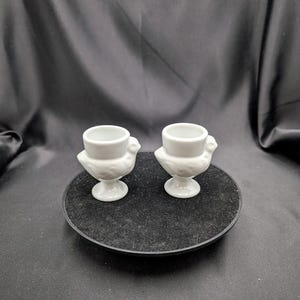 Op de afbeelding: Twee witte keramische eierdopjes, elk in de vorm van een kip, worden gepresenteerd op een zwart oppervlak. De eierdopjes hebben een kleine, ronde beker voor het ei en een voet. Het kippenontwerp omvat een gedetailleerd lichaam en hoofd.