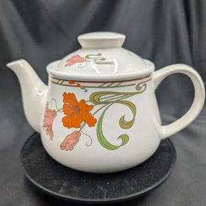 Puede incluir: Tetera de cerámica blanca con un diseño floral con flores naranjas, rosas y verdes. La tetera tiene una tapa y un asa.