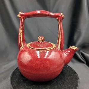 May include: A red ceramic teapot with a unique handle and a speckled glaze. The teapot has a small spout and a lid.