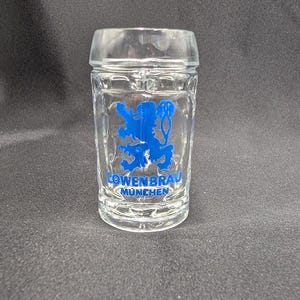 Könnte beinhalten: Klares Schnapsglas mit blauem Löwenemblem und der Aufschrift "LOWENBRAU MUNCHEN". Das Glas hat eine rechteckige Form mit dickem Boden und leicht ausgestelltem Rand. Für alkoholische Getränke.