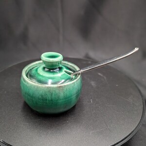 May include: A small, round, emerald green ceramic jar with a matching lid and a silver-toned metal spoon. The jar features a glossy, variegated glaze with darker green tones near the base and a small knob handle on the centre of the lid.