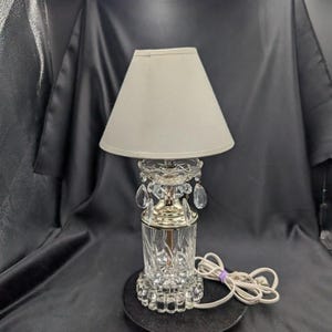 Puede incluir: Una lámpara de mesa de cristal con una pantalla beige. La lámpara presenta una base de cristal tallado, detalles metálicos plateados y adornos de cristal colgantes. La lámpara está sobre una base circular negra y tiene un cable de alimentación blanco.