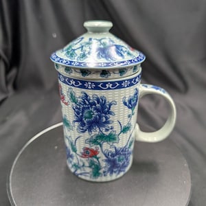 May include: A ceramic tea mug with a matching lid, featuring a white basket-weave texture background adorned with blue, green, and red floral patterns. The mug includes a curved handle and a blue decorative border around the rim and lid edge.