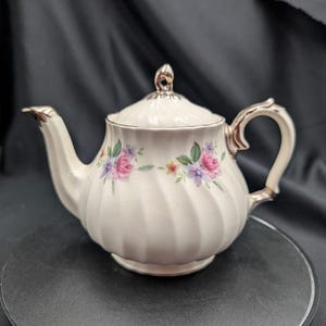 May include: A cream-coloured ceramic teapot with a swirling ribbed texture, featuring a delicate floral pattern of pink and purple roses with green leaves. The teapot includes a matching lid with a finial, a curved handle, and a spout with silver-toned metallic trim along the edges.