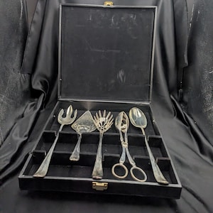 May include: A set of five silver-toned serving utensils arranged in a black velvet-lined presentation box. The collection includes a three-pronged fork, a triangular cake server, a slotted spoon, a pair of scissor-style tongs, and a solid spoon.