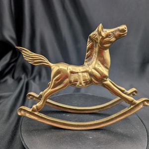 Könnte beinhalten: Eine Schaukelpferd-Figur aus poliertem Messing mit detaillierter Mähne, Sattel und fließendem Schweif. Das Pferd ist in einer Galopp-Pose dargestellt und auf zwei geschwungenen Kufen montiert, die eine Schaukelbewegung ermöglichen, mit metallischem Glanz.