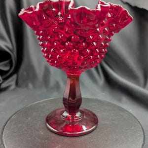 Op de afbeelding: Een dieprode glazen schaal met een hobnail-textuur over de kom en een gegolfde, geschulpte rand. Het object staat op een gladde, gefacetteerde voet die verbonden is met een ronde basis, wat een doorschijnende robijnrode kleur laat zien.
