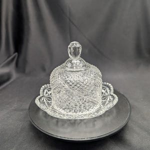 Op de afbeelding: Een heldere, geslepen glazen botervloot met een gewelfde deksel en een decoratieve, geschulpte basis. De deksel heeft een facetknop. De schaal is ontworpen met een diamant- en sterpatroon. De botervloot staat op een zwart oppervlak.
