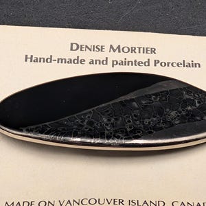 May include: Hand-made and painted porcelain brooch by Denise Mortier. The oval-shaped brooch features a glossy black surface with a textured, crackled design on one side. The other side is solid black. The brooch has a white border.
