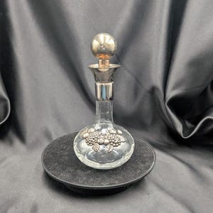 Peut inclure: Une carafe en verre transparent avec un bouchon argenté et un motif décoratif de grappes de raisin. Le bouchon est une boule ronde argentée. La carafe repose sur une surface noire et ronde sur un fond sombre.