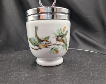 Pode incluir: Um cozedor de ovos de porcelana branca com uma ilustração pintada à mão de dois pequenos pássaros pousados em ramos de pinheiro. O recipiente tem uma tampa de metal prateado polido com uma argola no topo e uma base alargada.