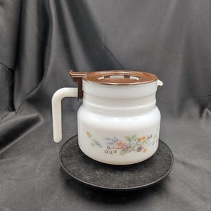 Op de afbeelding: Een witte glazen creamer met een bruin deksel en handvat. De creamer heeft een bloemenontwerp in pastelkleuren. Het staat op een zwarte, ronde onderzetter.