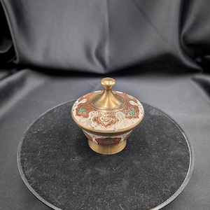 Peut inclure: Petit récipient décoratif en laiton avec couvercle. Le récipient présente des motifs complexes rouges, blancs et verts. Le couvercle et la base sont dorés. Le récipient est probablement utilisé pour ranger de petits objets.