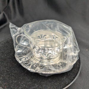 Può includere: Un set di tazza e piattino da tè argentati, con motivi ornati, è avvolto in plastica trasparente. Il set è esposto su una superficie scura e rotonda, creando un contrasto con il metallo lucido.