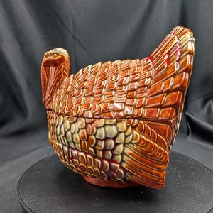 May include: A ceramic turkey-shaped serving dish with a lid. The dish is mainly brown with orange and cream accents, and features detailed feather patterns. The turkey has a long neck and a detailed head. The dish is likely for autumn or Thanksgiving.
