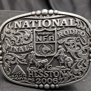 Op de afbeelding: Een ovale zilverkleurige metalen riemgesp met in reliëf de tekst NATIONAL FINALS RODEO, NFR, Wrangler, HESSTON 2006 en AGCO PARTS. Het ontwerp bevat gedetailleerde reliëfsculpturen van twee figuren te paard en sierlijk krulwerk.