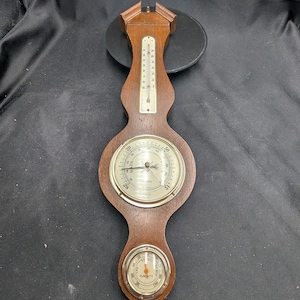 Puede incluir: Una estación meteorológica de madera con termómetro, barómetro e higrómetro. El termómetro está en la parte superior, el barómetro en el medio y el higrómetro en la parte inferior. Los indicadores tienen una esfera plateada con marcas negras y un marco de madera.