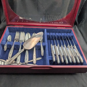 Könnte beinhalten: Ein Besteckset in einer Holzkiste mit rotem Rahmen und Glasdeckel. Das Besteck umfasst Löffel, Gabeln, Messer und einen Tortenheber, alles angeordnet auf einem blauen Samtinnenraum.