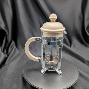 May include: A glass French press coffee maker featuring a beige lid with a matching beige handle. The carafe is encased in a polished chrome metal frame with four legs and includes a stainless steel plunger mechanism with a fine mesh filter.