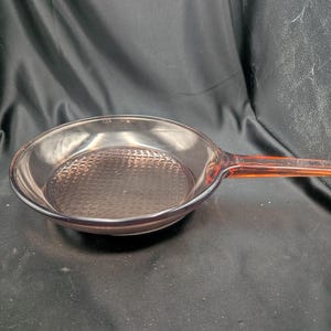 May include: A translucent, light purple glass frying pan with a round, shallow basin and a flat, extended handle. The interior base of the pan features a distinct, raised grid-like texture pattern, while the exterior surface remains smooth.