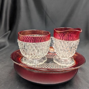 May include: A vintage glass creamer and sugar bowl set with a ruby red rim and diamond pattern. The set includes a small creamer jug, a sugar bowl, and a matching serving tray. The set is made of clear glass.