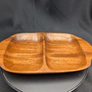 Könnte beinhalten: Ein Holzserviertablett mit zwei Fächern. Das Tablett besteht aus warm getöntem Holz mit sichtbaren Maserungsmustern. Das oval geformte Tablett hat abgerundete Kanten und ist zum Servieren von Snacks oder Vorspeisen konzipiert. Der Hintergrund ist dunkel.