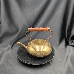 Pode incluir: Uma pequena chaleira de latão antiga com uma alça de madeira. A chaleira tem um corpo arredondado, um bico e um design decorativo gravado. A alça é feita de madeira e é presa à chaleira com suportes de latão. A chaleira está em um prato preto.