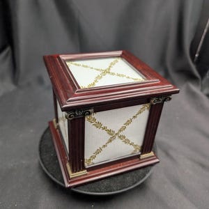May include: A rectangular, dark wood box with a glass top and sides. The box features gold-coloured accents and a white fabric panel with a gold leaf pattern. The box is set on a black surface.