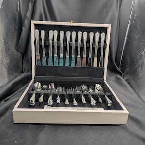 May include: A full set of stainless steel cutlery presented in a light-coloured wooden case. The set includes knives, forks, and spoons, arranged in a black velvet-lined interior. The case is open, displaying the cutlery.