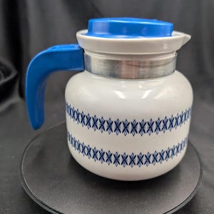 May include: White ceramic coffee pot with a blue lid and handle. The pot has a silver band around the middle and a blue and white geometric pattern. The pot is sitting on a black surface.