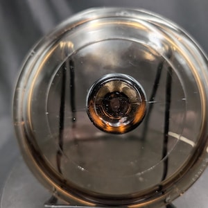 Pode incluir: Uma tampa de vidro circular, translúcida e de cor fumê, com um puxador central redondo de cor castanho-escuro. A tampa apresenta uma superfície lisa e curva com sulcos circulares concêntricos e uma borda exterior ligeiramente elevada.