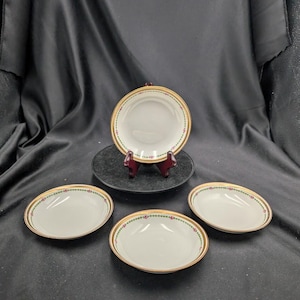 May include: Four white porcelain dishes with gold rims and floral detailing. One dish is displayed on a black stand. The oval-shaped dishes are suitable for serving appetizers or desserts. The dishes measure approximately 15 cm in length.